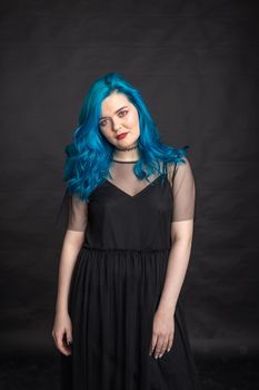 People and fashion concept - Woman with blue long hair dressed in black dress posing over black background.