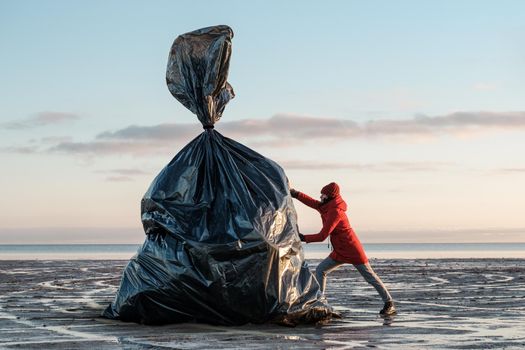 A woman pushes a huge black trash bag on the shore. Environmental pollution by plastic. Recyclable packaging.