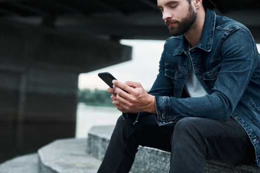 Outdoors leisure. Young stylish man sitting on stairs on city street browsing internet on smartphone concentrated close-up