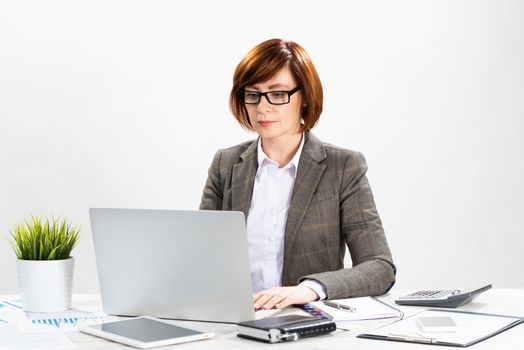 Attractive middle aged businesswoman working with computer in office. Business lady sitting at desk on white background. Successful corporate woman in suit and glasses posing at her workplace.