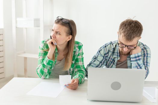 Young husband and wife are upset about numerous checks for payments and purchases for repairs while sitting at the table with a laptop. The concept of low living standards and savings. Copyspace