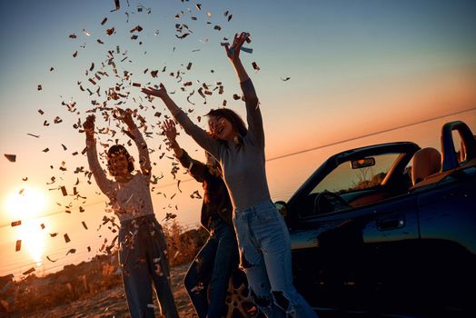 Cheerful young three women are dancing near the cabriolet. They throw up falling leaves. Beautiful sunset at the background