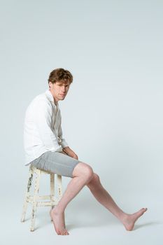 Vertical image. Sitting half turned on the rusty chair full length shot of young handsome young man wearing white shirt and grey shorts looking at camera isolated on white background.