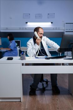 Medic using telephone call for remote communication overtime. Doctor talking on landline phone in office for healthcare and checkup appointment, working late. Man on phone call
