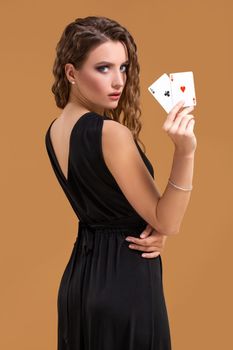 Beautiful brown-haired woman in black dress holding two aces as a sign for poker game, gambling and casino on a beige background. Studio shot
