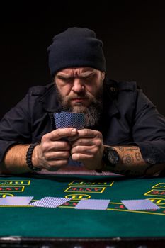 Serious bearded man in a dark blue hat sitting at poker table and holding cards isolated on black. The concept of gambling