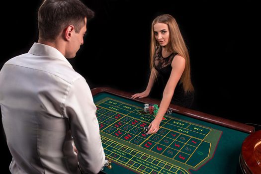 A close-up on the back of the croupier in a white shirt, image of green casino table with roulette and chips, a rich woman betting of gambling in the background. Casino. Gambling. Roulette. Betting