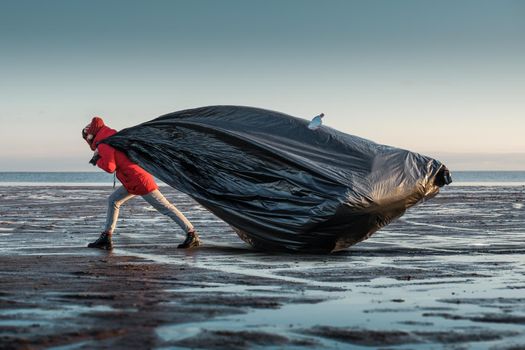 A woman drags a huge black trash bag along the shore. Environmental pollution by plastic. Recyclable packaging.