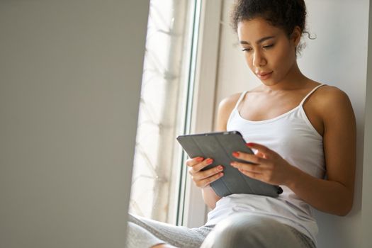 Focused young mixed race woman reading a book using tablet pc, feeling cozy while sitting alone by the window at home. Lifestyle, leisure concept