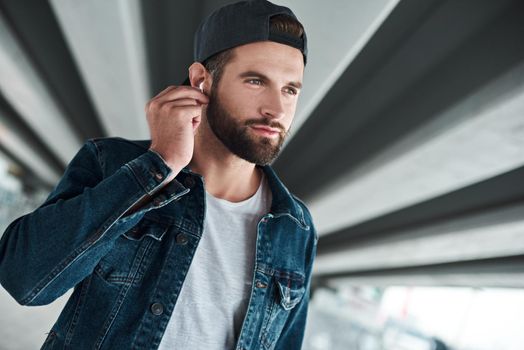 Outdoors leisure. Young stylish man walking on city street putting on wireless earphones listening to music smiling joyful close-up