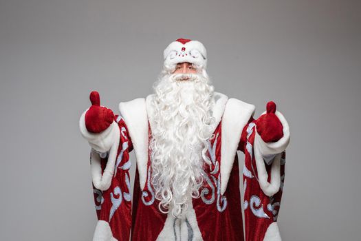 Stock photo of white-bearded Father Frost in red and white hat and traditional costume with red mittens showing both thumbs up.