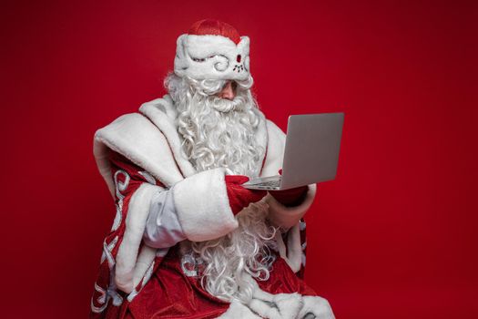 Senior Santa Claus with long white beard wearing traditional fancy costume holding laptop in both hands and reading emails from children. Isolated on red.