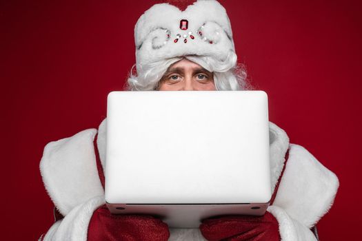 Stock photo of curious Santa Claus in fancy hat and traditional costume holding laptop in hands and looking at camera. Cutout on red background.