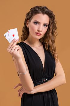 Beautiful brown-haired woman in black dress holding two aces as a sign for poker game, gambling and casino on a beige background. Studio shot