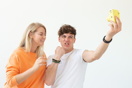 Funny crazy young couple blonde girl and a hipster guy taking a selfie on a vintage yellow film camera posing on a white background. Concept of photography hobby. Advertising space