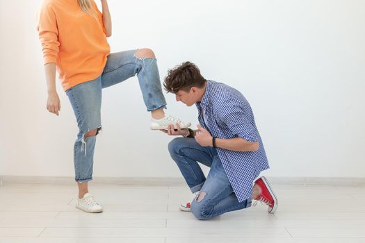 Young man is kneeling and reverently tying shoelaces to his domineering unidentified woman posing on a white background. Concept of dominant relationships