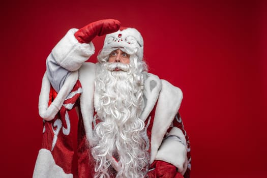 Studio portrait of white-bearded Father Frost in traditional fancy clothes holding hand in red mitten at his hat looking away and searching something. Isolate on red.