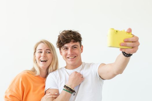 Funny crazy young couple blonde girl and a hipster guy taking a selfie on a vintage yellow film camera posing on a white background. Concept of photography hobby. Advertising space