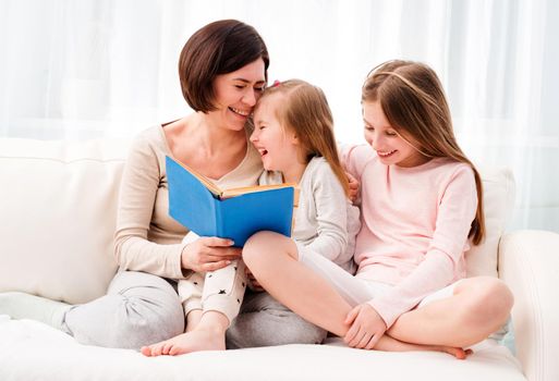 Mom and her two little daughters reading children's book in the living room. Mom and daughters sitting on the coach and reaading