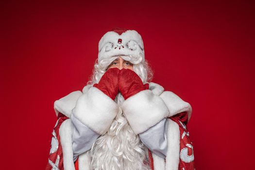 Stock photo of Santa Claus in fancy traditional costume covering mouth with both hands worn in red mittens. Silence concept.