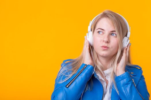 Portrait of pretty young girl blonde female student holding smartphone with blue leather jacket headphones posing on a yellow background. Concept of listening to online radio and music subscription