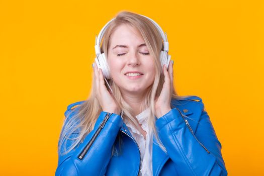 Portrait of pretty young girl blonde female student holding smartphone with blue leather jacket headphones posing on a yellow background. Concept of listening to online radio and music subscription