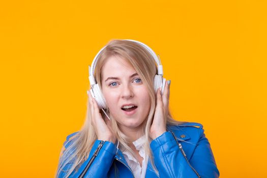 Portrait of pretty young girl blonde female student holding smartphone with blue leather jacket headphones posing on a yellow background. Concept of listening to online radio and music subscription