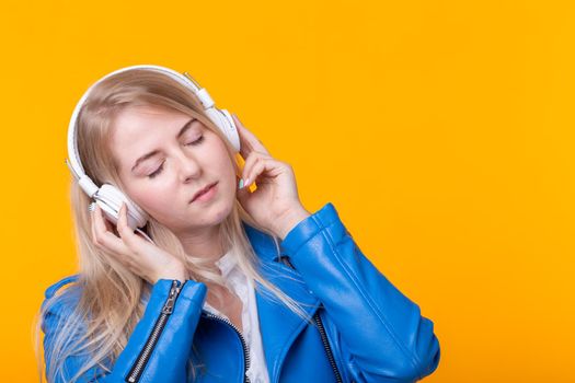 Portrait of pretty young girl blonde female student holding smartphone with blue leather jacket headphones posing on a yellow background. Concept of listening to online radio and music subscription