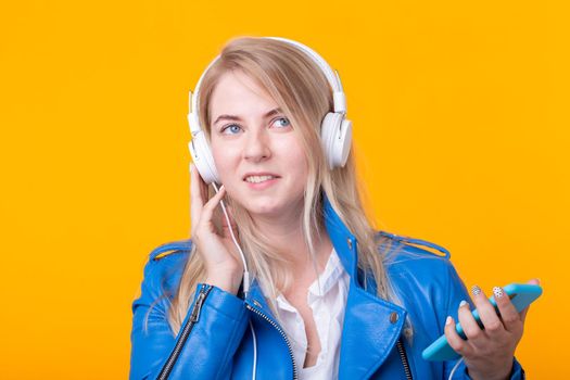 Portrait of pretty young girl blonde female student holding smartphone with blue leather jacket headphones posing on a yellow background. Concept of listening to online radio and music subscription