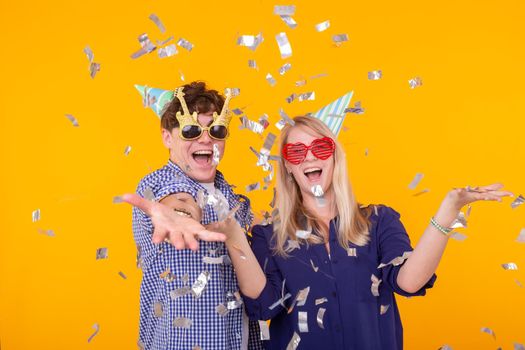 Young cheerful positive couple of funny glasses and a paper cap rejoice and shout on a yellow background with flying confetti. Conception of a holiday and fun