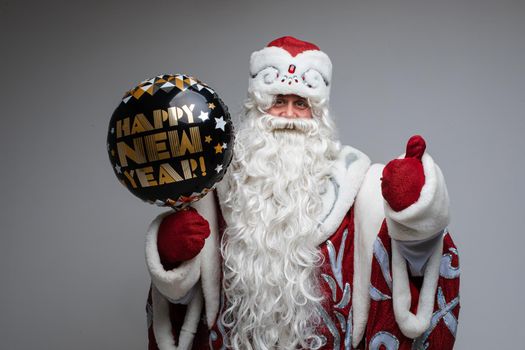 Happy Santa Claus in traditional fancy costume with inflatable black ballon with golden text Happy New Year showing thumb up.