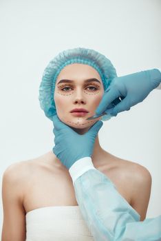 Anti aging treatment. Beautiful young woman in blue medical hat with dashed lines on her face having cosmetic face surgery plastic surgery. Plastic surgeon in blue gloves holding scalpel. Beauty concept.