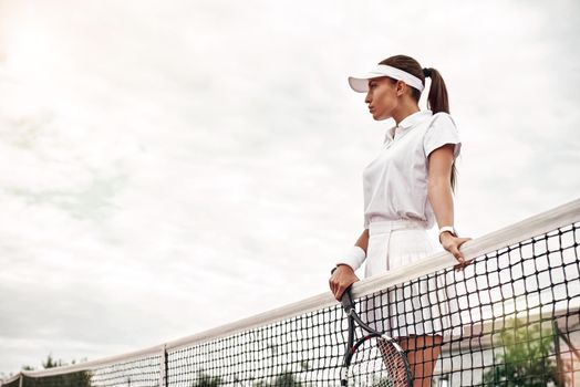 Female tennis player in white sportswear with a racket in her hand stands on a court, looking away. Healthy lifestyle