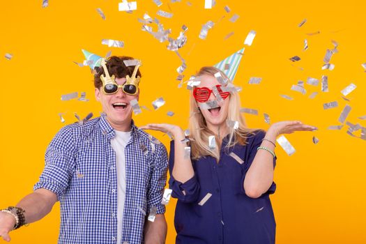 Young cheerful positive couple of funny glasses and a paper cap rejoice and shout on a yellow background with flying confetti. Conception of a holiday and fun