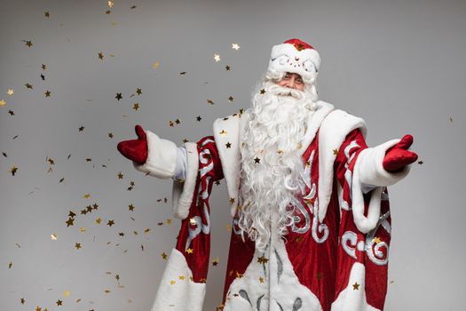 Stock photo of jolly Father Frost in festive clothing with long white beard outstretching his arms and smiling at camera under flying golden confetti. Isolated on grey background.