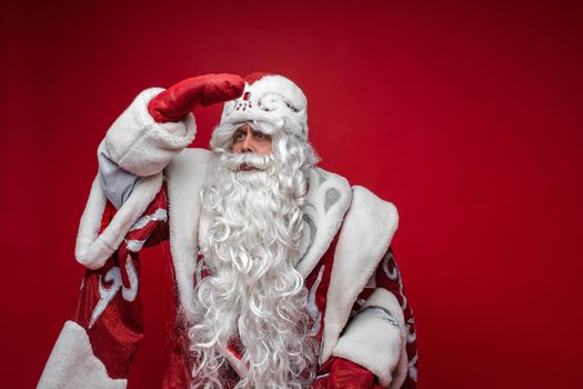 Festive Santa Claus look into the distance with hand on forehead on red background, copy space for holiday advertising. High quality photo