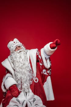 Studio photo of white-bearded Santa in red and white fancy costume with long white beard pointing away with his arm. Isolate on red background. Showing the direction.