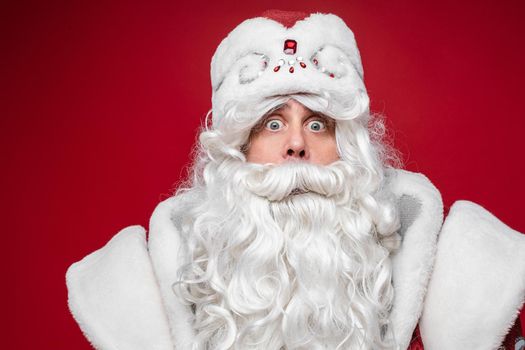 Surprised shocked Santa Claus, wondering senior male with gray beard, close up studio portrait on red background. High quality photo