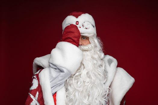 Stock photo of serious Santa Claus in traditional fancy costume covering one half of his face with hand in red mitten.