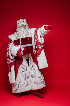 santa claus with long white beard shows something with his hand and holds a gift, picture isolated on red background