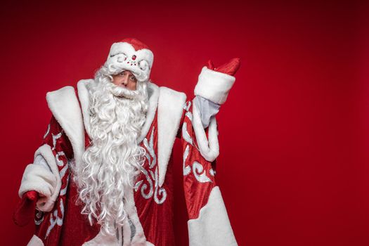 Santa Claus elderly male in festive xmas costume point by hand to copy space on red background. High quality photo