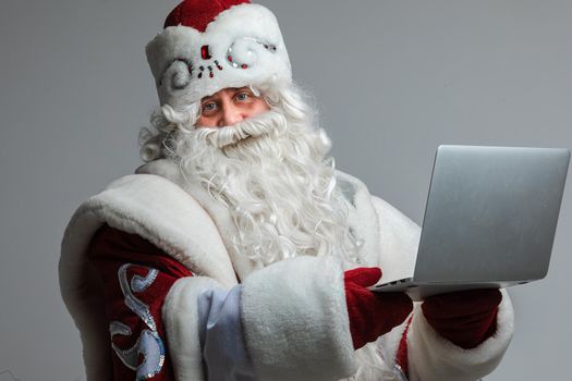 Stock photo of smiling happy Father Frost with white long beard in festive winter hat and traditional coat holding laptop and looking at camera.