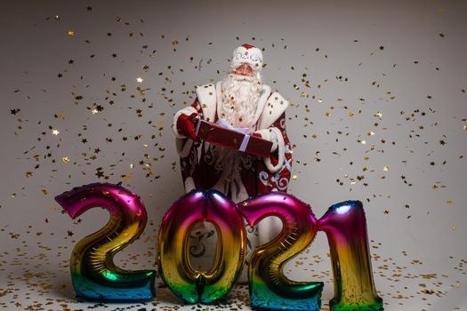 Stock photo of jolly Father Frost in festive clothing with long white beard outstretching his arms and smiling at camera under flying golden confetti. Isolated on grey background.