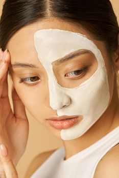 Close up shot of young woman with facial mask applied on half of her face receiving spa treatments, posing for camera isolated over beige background. Beauty, skincare concept