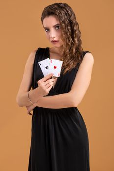 Beautiful brown-haired woman in black dress holding two aces as a sign for poker game, gambling and casino on a beige background. Studio shot