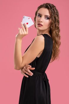 Young woman in black dress holding two aces in hand against on pink background. Studio shot
