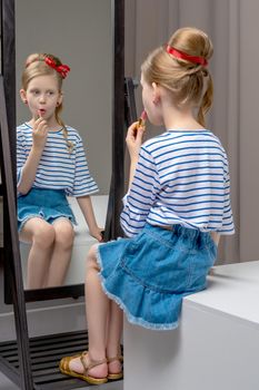 A nice little girl near the big mirror paints her lips with lipstick. The concept of childhood, style and fashion.