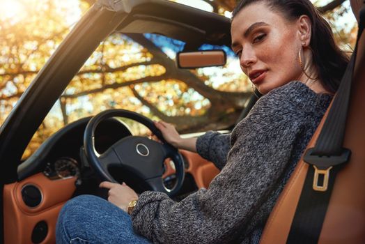 Sexy young woman in jeans and in a grey sweater sits in front seat of a cabriolet. Autumn season