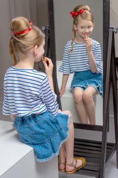 A nice little girl near the big mirror paints her lips with lipstick. The concept of childhood, style and fashion.