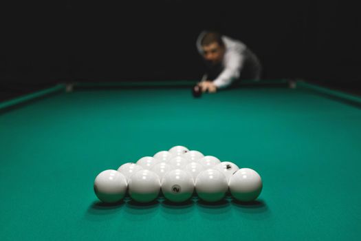 Focus on the pile of white billiard balls and hand of a man in a white shirt aiming for a kiy. Draw of the right of the first blow.